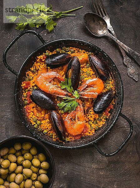 Paella in schwarzer Pfanne mit Reis  Krabben  Muscheln  Tintenfisch und Fleisch  Schüssel mit Oliven und Vintage Besteck. Paella mit Meeresfrüchten  traditionelles spanisches Gericht. Paella auf rustikalem Tisch. Selektiver Fokus. Ansicht von oben