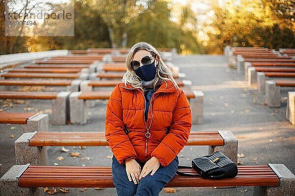 Blonde Frau in modischer Oberbekleidung und Stoffmaske blickt in die Kamera  während sie auf einer Parkbank während einer Pandemie sitzt