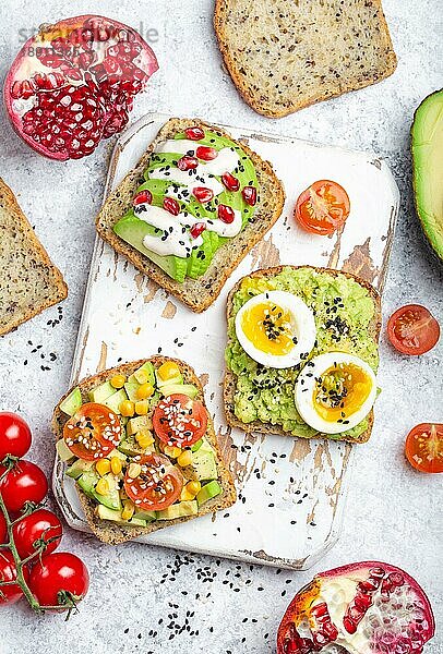 Avocado-Toasts mit Ei  Tomaten  Gewürze auf weißem Holz rustikalen Schneidebrett  Stein Hintergrund. Zutaten für gesunde Frühstücks-Avocado-Sandwiches mit verschiedenen Belägen  Draufsicht  Nahaufnahme