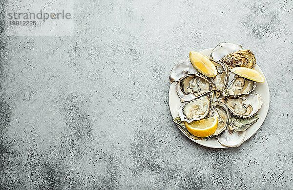 Nahaufnahme von einem halben Dutzend frisch geöffneter Austern und Muscheln mit Zitronenscheiben auf einem Teller  Draufsicht  grauer rustikaler Betonhintergrund  Platz für Text. Delikatesse Austern für Snack  Mittagessen