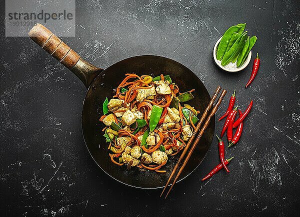 Gebratenes Huhn mit Gemüse in einer alten  rustikalen Wokpfanne  Stäbchen auf schwarzem Steinhintergrund  Nahaufnahme  Draufsicht. Traditionelle asiatische/thailändische Mahlzeit