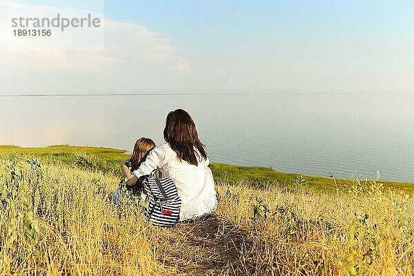 Rückenansicht einer nicht erkennbaren Frau  die ein Mädchen umarmt  während sie auf getrocknetem Gras sitzt und eine malerische Meereslandschaft an einem Sommerwochenende bewundert