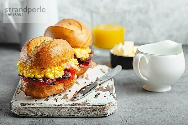 Frühstückssandwiches mit Rührei  Speck  Käse  Tomaten auf weißem Holzbrett  Glas mit frischem Orangensaft  weißer Hintergrund. Frühstück machen Konzept  selektiver Fokus