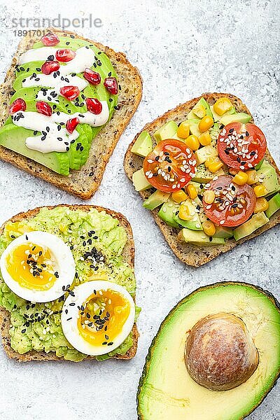 Avocado-Toasts mit Ei  Tomaten  Gewürzen und einer halben ganzen Avocado auf weißem Steinhintergrund. Gesunde Frühstücks-Avocado-Sandwiches mit verschiedenen Belägen  Draufsicht  Nahaufnahme