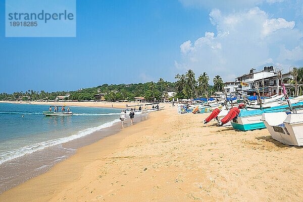 Am Strand eines der wichtigsten Touristenzentren im Südwesten Sri Lankas. Touristen nehmen ein Sonnenbad und treiben Wassersport. Auslegerboote und traditionelle Fischerboote am Strand und vor Anker