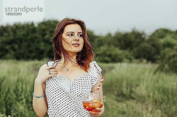 Junge Frau trinkt Alkoholcocktail mit Orange auf unscharfem Hintergrund der Natur