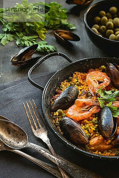 Paella in schwarzer Pfanne mit Reis  Krabben  Muscheln  Tintenfisch und Fleisch  Schüssel mit Oliven und Vintage-Besteck. Paella mit Meeresfrüchten  traditionelles spanisches Gericht. Paella auf einem rustikalen schwarzen Holztisch. Selektiver Fokus