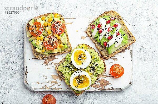 Avocado-Toasts mit Ei  Tomaten  Gewürze über weiße Holz rustikal Schneidebrett auf Stein Hintergrund. Herstellung gesunder Frühstücks-Avocado-Sandwiches mit verschiedenen Belägen  Draufsicht  Nahaufnahme