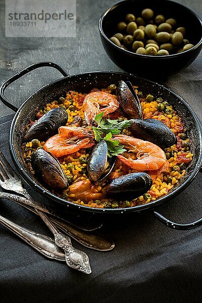 Paella in schwarzer Pfanne mit Reis  Krabben  Muscheln  Tintenfisch und Fleisch  Schüssel mit Oliven und Vintage-Besteck. Paella mit Meeresfrüchten  traditionelles spanisches Gericht. Paella auf einem rustikalen schwarzen Holztisch. Selektiver Fokus