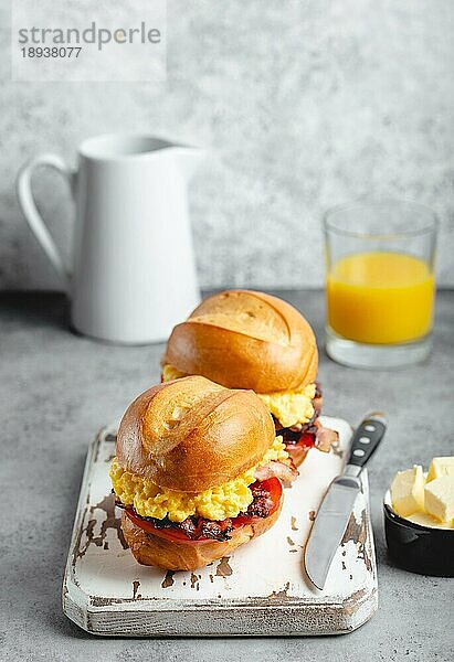 Frühstückssandwiches mit Rührei  Speck  Käse  Tomaten auf weißem Holzbrett  Glas mit frischem Orangensaft  weißer Hintergrund. Frühstück machen Konzept  selektiver Fokus