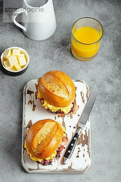 Draufsicht auf Frühstückssandwiches mit Rührei  Speck  Käse  Tomaten auf weißem Holzbrett  Glas mit frischem Orangensaft  weißer Hintergrund. Frühstück machen Konzept  selektiver Fokus  Nahaufnahme