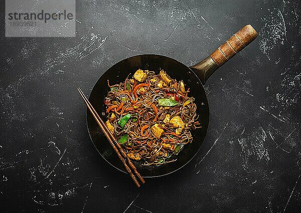 Rührbraten Soba-Nudeln mit Huhn  Gemüse in alter rustikaler Wok-Pfanne  Stäbchen auf schwarzem Stein-Hintergrund  Nahaufnahme  Draufsicht. Traditionelle asiatische/thailändische Mahlzeit