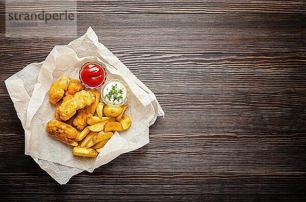 Britische traditionelle Fast-Food-Fisch und Chips mit verschiedenen Dips zur Auswahl  auf Papier  rustikaler Holzhintergrund  Draufsicht  Platz für Text. Gebackener gebratener Fisch  Kartoffelchips  Tartar und Ketchup-Sauce