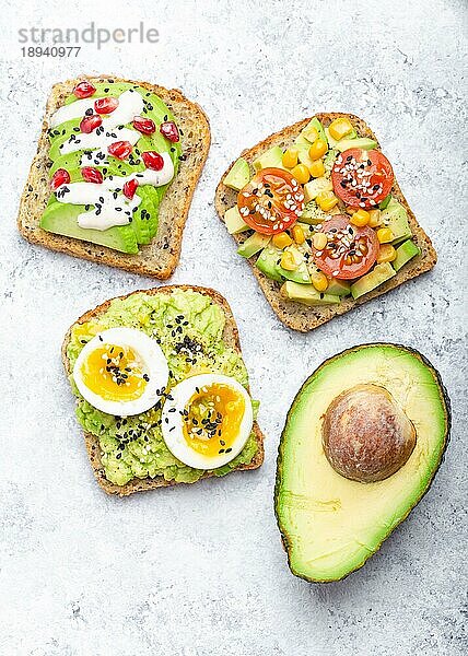 Avocado-Toasts mit Ei  Tomaten  Gewürzen und einer halben ganzen Avocado auf weißem Steinhintergrund. Gesunde Frühstücks-Avocado-Sandwiches mit verschiedenen Belägen  Draufsicht  Nahaufnahme