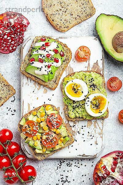 Avocado-Toasts mit Ei  Tomaten  Gewürze auf weißem Holz rustikalen Schneidebrett  Stein Hintergrund. Zutaten für gesunde Frühstücks-Avocado-Sandwiches mit verschiedenen Belägen  Draufsicht  Nahaufnahme