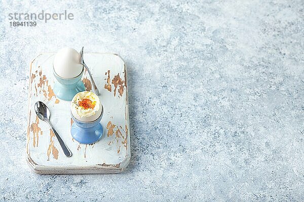 Frische weich gekochte Eier in Ständern  Löffel  Holzkochbrett  weißer Beton rustikalen Hintergrund. Ansicht von oben. Weiche Eier  gesundes Frühstück. Platz für Text. Selektiver Fokus. Eiweiß-Fitness-Frühstück