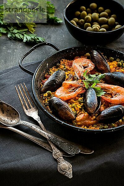 Paella in schwarzer Pfanne mit Reis  Krabben  Muscheln  Tintenfisch und Fleisch  Schale mit Oliven und Vintage Besteck. Paella mit Meeresfrüchten  traditionelles spanisches Gericht. Paella auf einem rustikalen schwarzen Holztisch. Selektiver Fokus