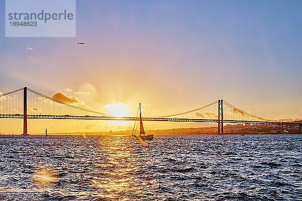 Blick auf die Brücke 25 de Abril  das berühmte touristische Wahrzeichen von Lissabon  die Lisboa und Almada auf der Halbinsel Setubal über den Fluss Tejo mit der Silhouette einer Touristenyacht bei Sonnenuntergang und einem fliegenden Flugzeug. Lissabon  Portugal  Europa