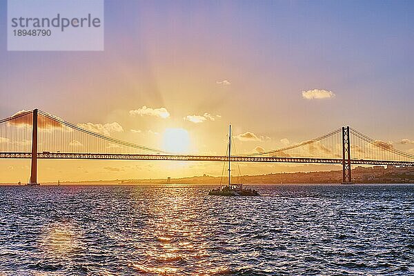 Blick auf die Brücke 25 de Abril  das berühmte touristische Wahrzeichen von Lissabon  das Lisboa und Almada auf der Halbinsel Setubal verbindet  über den Fluss Tejo mit der Silhouette einer Touristenyacht bei Sonnenuntergang. Lissabon  Portugal  Europa