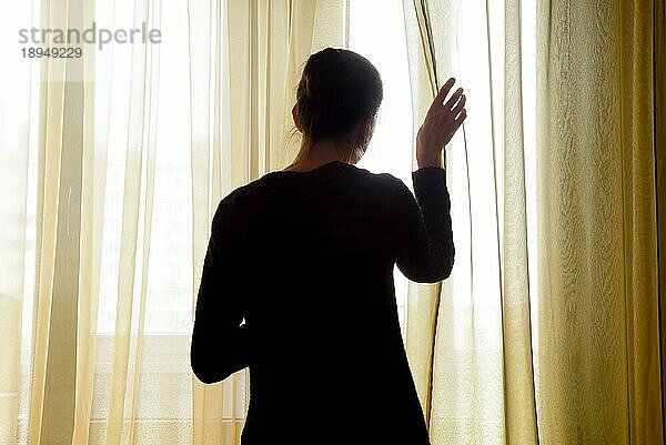 Eine Frau in Schwarz schiebt den Vorhang beiseite  um durch das Fenster hinauszuschauen
