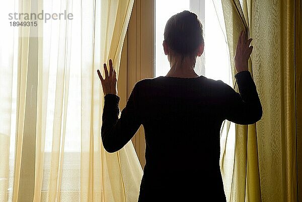 Eine Frau in Schwarz schiebt den Vorhang beiseite  um durch das Fenster hinauszuschauen