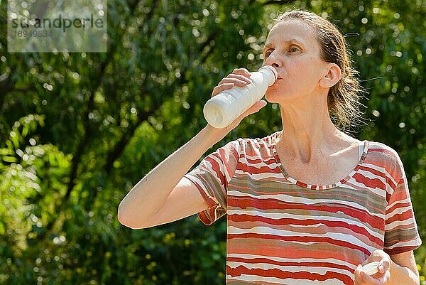 Eine ältere Frau trinkt Wasser oder Joghurt  um ihren Durst nach einem langen Lauf im Wald zu stillen