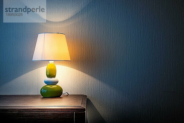 Kleine gelbe  graue und grüne Lampe auf einer hölzernen Kommode oder einem Nachttisch  die die mit Tapete bedeckte Wand nachts oder spät abends beleuchtet. Weiche und warme Innen Stimmung