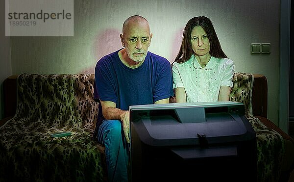 Ein erwachsener Mann und eine erwachsene Frau sitzen traurig auf einer Couch und sehen fern. Sie sehen einen traurigen Film oder hören schlechte Nachrichten  die sie ein wenig unglücklich und deprimiert machen