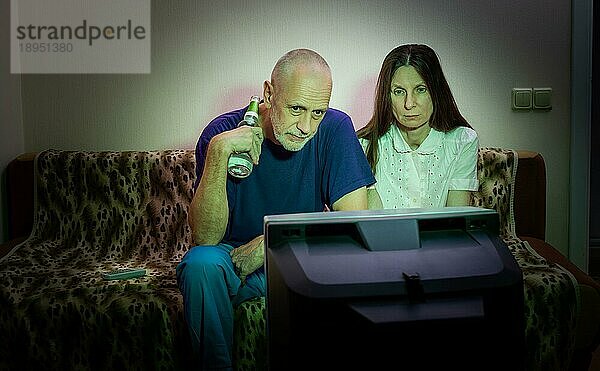 Ein Mann und eine Frau mittleren Alters sitzen traurig auf einer Couch und sehen fern. Sie sehen einen traurigen Film oder hören schlechte Nachrichten  die sie ein wenig unglücklich und deprimiert machen