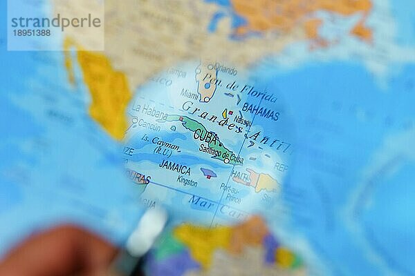Hand eines Mannes mit einem Vergrößerungsglas  der Kuba auf einer Weltkarte betrachtet