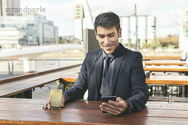 Geschäftsmann sitzt an einer Bar im Freien  trinkt Limonade und schaut lächelnd auf sein Telefon