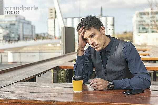 Geschäftsmann sitzt in einer Bar im Freien und trinkt einen Kaffee und hält sich mit besorgter Miene den Kopf