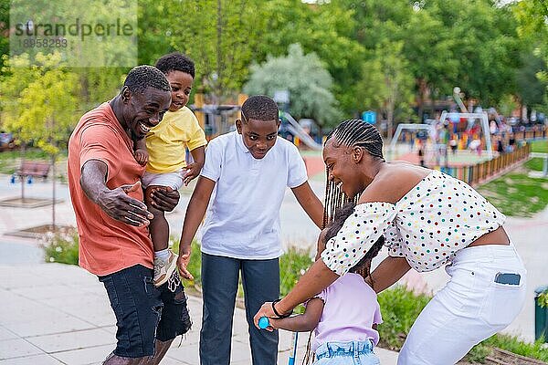 Afrikanische schwarze ethnische Familie mit Kindern auf dem Spielplatz  die sich mit dem Fahrrad umarmen