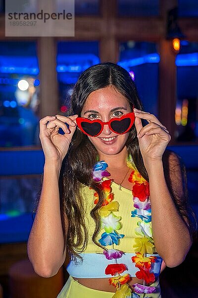 Porträt einer attraktiven lateinamerikanischen Frau  die bei einer Nachtparty in einer Diskothek lächelt und eine Brille mit Herzen trägt