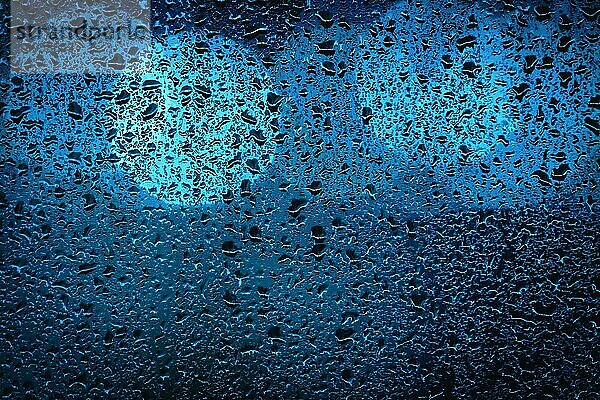 Blaue Bokehlichter hinter einem naßen Fenster an einem regnerischen Abend in einer Großstadt