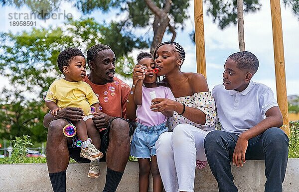Schwarzafrikanische Familie mit Kindern auf dem Spielplatz  die sich beim Blasen von Seifenblasen neben den Bäumen im Park vergnügen