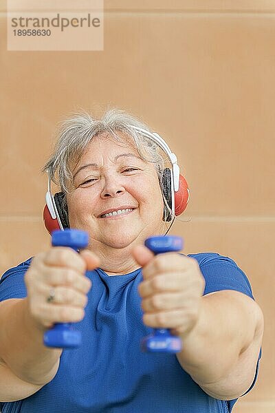 Frontalansicht einer fettleibigen weißhaarigen älteren Frau  die mit Hanteln und Kopfhörern trainiert und Musik hört. lächelnd