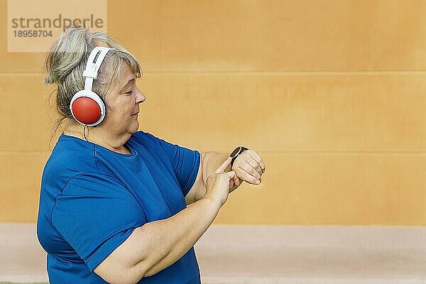 Ältere Frau mit Übergewicht  Sportkleidung und Kopfhörern konsultiert ihre Smartwatch beim Training im Park
