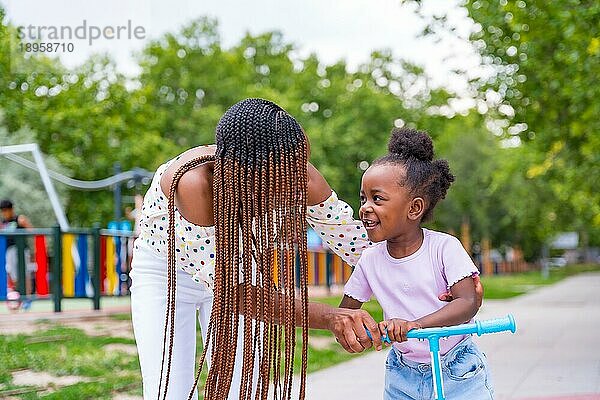 Afrikanische schwarze ethnische Mutter hat Spaß mit ihrer Tochter auf dem Spielplatz mit dem Skateboard gehen