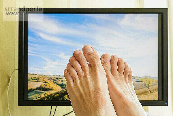 Die Füße eines Mannes vor einem Computermonitor mit einer schönen sonnigen italienischen Landschaft mit bewölktem Himmel  um den Wunsch nach Erholung im Urlaub zu symbolisieren