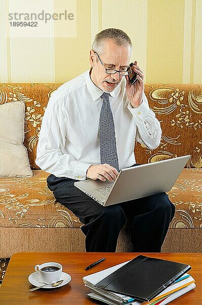 Ein gutaussehender älterer Geschäftsmann  der zu Hause am Computer arbeitet  mit seinem Mobiltelefon einen Partner anruft und bereit ist  eine heiße Tasse schwarzen Kaffee zu trinken