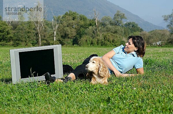 Glückliche Frau mit ihrem Hund  die auf dem grünen Gras liegt und an einem sonnigen Tag auf dem Feld fernsieht