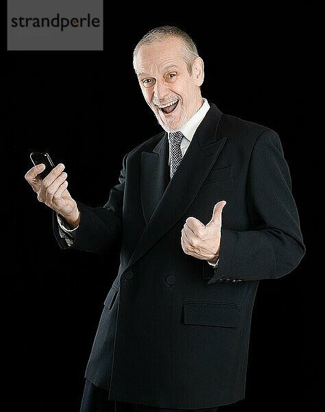 Glücklicher Geschäftsmann im schwarzen Anzug  lächelnd und SMS auf dem Handy lesend  mit Daumen nach oben  auf schwarzem Hintergrund