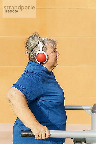 Profilansicht einer aktiven  übergewichtigen älteren Frau in Sportkleidung  die in einem Fitnessstudio im Park an einem Gerät trainiert  während sie über Kopfhörer Musik hört