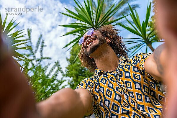 Afro haired Mann im Sommerurlaub neben einigen Palmen neben dem Strand ein Selfie mit beiden Händen lächelnd zu nehmen. Reisen und Tourismus Konzept