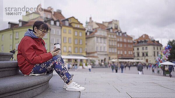 Ältere Dame sitzt auf den Stufen  trinkt Kaffee und benutzt ein Smartphone im historischen Zentrum einer alten europäischen Stadt. Schlossplatz  Altstadt von Warschau