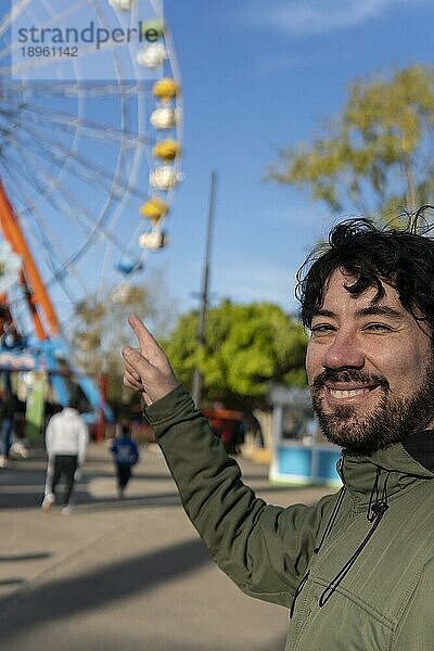 Porträt eines Latinomannes in einem Vergnügungspark  der glücklich mit dem Riesenrad im Hintergrund posiert