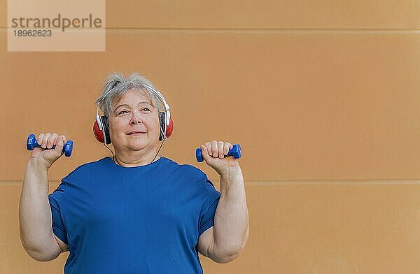Weißhaarige fettleibige ältere Frau  die mit Hanteln und Kopfhörern trainiert und Musik zur Gewichtsabnahme hört  vororangefarbenem Hintergrund und Kopierraum