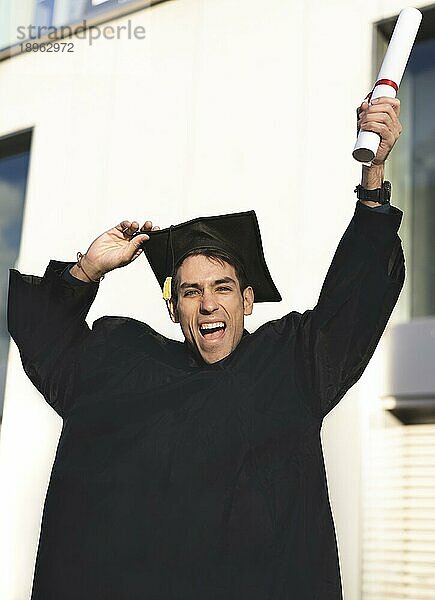 Fröhlich lächelnder Absolvent  der einen Talar trägt und sein Diplom hochhält