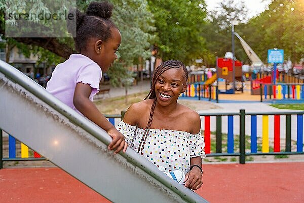 Afrikanische schwarze ethnische Mutter hat Spaß mit ihrer Tochter in der Quietsche des Spielplatzes des Stadtparks in den Sonnenuntergang